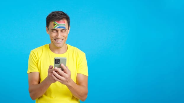 South african supporter with the flag of South Africa painted on his face, looking at his mobile phone smiling