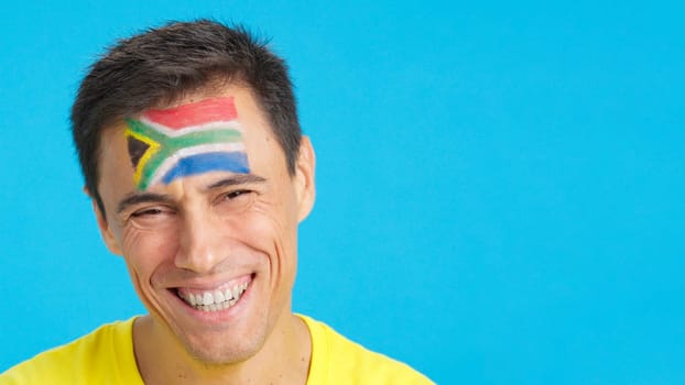 Close up of a caucasian man with a south african flag painted on the face smiling at camera