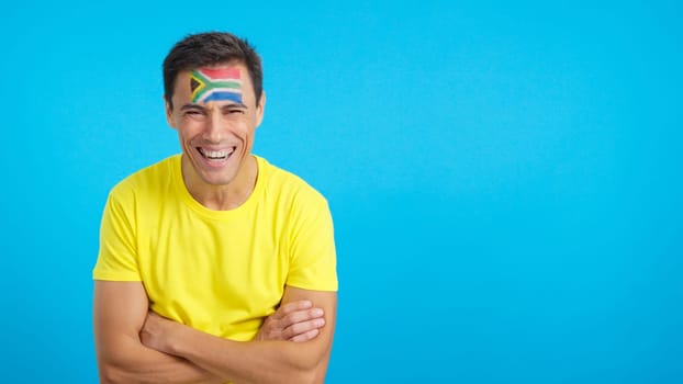 Man standing with south african flag painted on face smiling with arms crossed