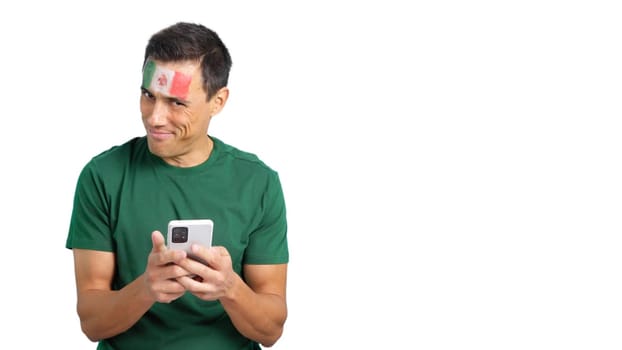 Mexican supporter with the flag of Mexico painted on his face, looking at his mobile phone smiling