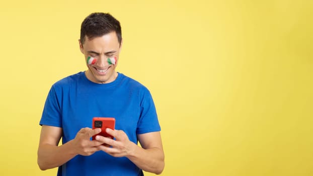 Italian man with the flag of Spain painted on his face, looking at his mobile phone smiling