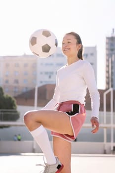 smiling female soccer player kicking the ball with her knee on an urban football court, concept of sport and active lifestyle