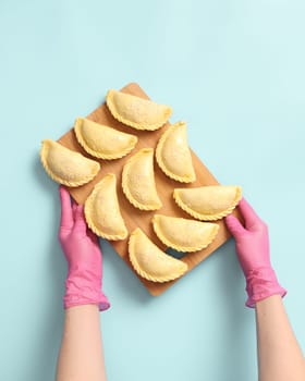 Top view on woman baker's hands in a pink rubber gloves with wooden board with raw pies on blue background, frozen semi-finished products.