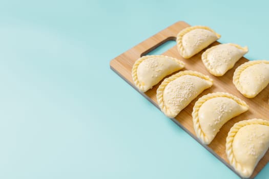 Raw samosas with sesame on a wooden on board on blue table ready for baking. Traditional Central Asian cuisine, frozen semi-finished products, copy space.
