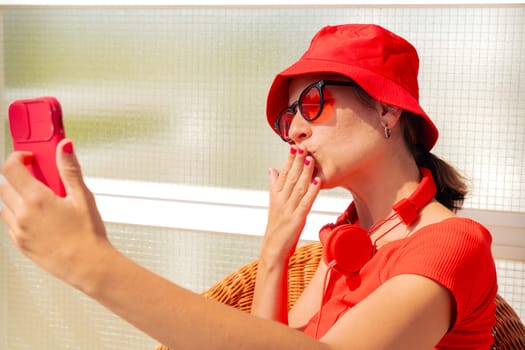Stylish young woman smiling doing a video call with smartphone wearing glasses and red hat. Rait of a beautiful Caucasian woman wearing red hat and glasses smiling looking at the camera.
