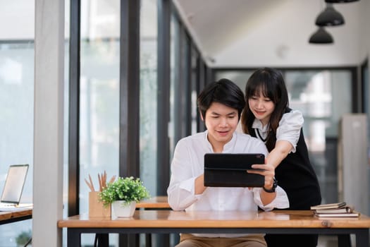 Two university students consulting together on homework in a modern study space, using a digital tablet and surrounded by books.