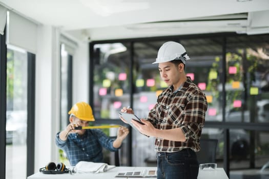 Two male engineer in a modern office, wearing safety helmets, engaged in project planning and development with measuring tools and sticky notes in the background.