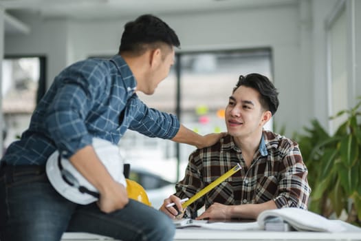 Two male engineers in a modern office, discussing blueprints and measurements, wearing casual plaid shirts, and engaging in a productive conversation.