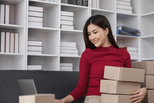 Female entrepreneur packing products for online sales, working on laptop in modern office with shelves.