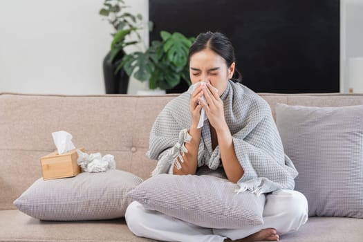 A woman with a fever sits on a sofa, wrapped in a blanket, holding a tissue, and looking unwell in a modern living room setting.