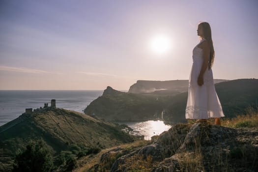 A woman stands on a rocky hill overlooking the ocean. She is wearing a white dress and she is enjoying the view. The scene is serene and peaceful, with the sun shining brightly in the background