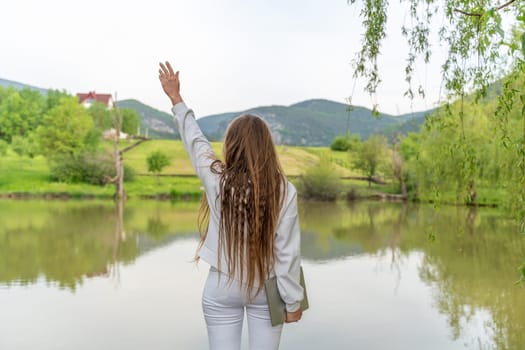 A woman with long hair is standing by a lake, holding her hand up in the air. The scene is peaceful and serene, with the woman's pose and the calm water creating a sense of tranquility