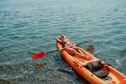 A woman is laying on a kayak in the water. The kayak is orange and has a paddle on it. The woman is wearing a red hat