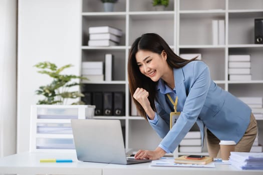 Young female accountant in a modern office, smiling and celebrating success while working on a laptop with documents.