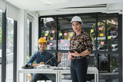 Two male engineer in a modern office, working with blueprints and a digital tablet, wearing safety helmet and casual attire, surrounded by sticky notes.