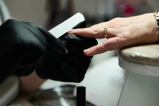 A nail technician wearing black gloves files a client's nails in a beauty salon, showcasing meticulous care and attention to detail.