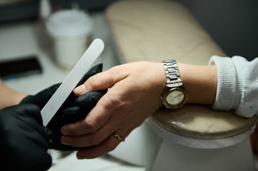 A close-up image of a manicure service in a beauty salon. An aesthetician with gloves works on a client's nails using a nail file.