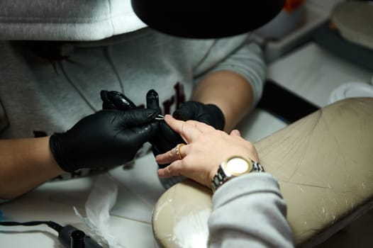 A professional nail technician wearing black gloves meticulously working on a client's manicure at a beauty salon, focusing on precision and detail.