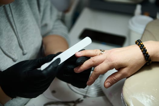 Close-up of a manicurist filing nails at a beauty salon. Focus on hands, black gloves, and nail file. Professional manicure service.