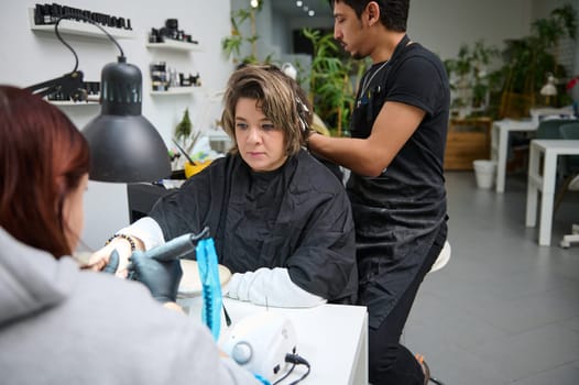 A woman enjoys getting her hair styled and nails done at a beauty salon, attended to by two professionals, showcasing a relaxing day in a beauty environment.