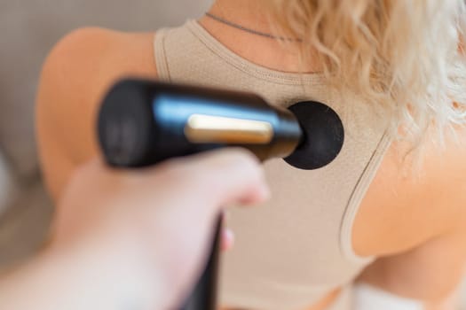 Girl at home holding a therapeutic percussion vibrating massage gun in her hand and massaging her back muscles Sports recovery concept after a workout. . High quality photo