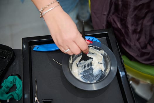 Hairdresser's hand preparing hair dye in a bowl using a brush in a beauty salon. Essential salon tools are visible on the table.