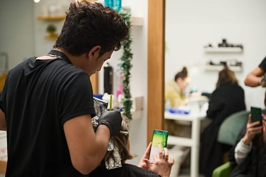 Hair stylist applying color to client's hair in a busy beauty salon. Clients are seen using their phones while waiting.