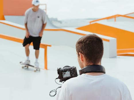 Videographer filming skateboarding in skatepark. Professional cameraman - covering on event with video. Cameraman behind scene of man on skateboard in front of camera
