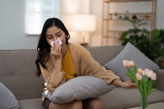 Young woman in a yellow sweater sneezing into a tissue due to pollen allergy, holding a vase of flowers while sitting on a couch