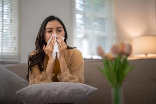 Young woman sitting on a couch sneezing into a tissue due to pollen allergy, with a vase of flowers in the foreground