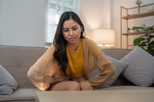 A young woman sitting on a couch appears to be in discomfort, clutching her stomach with a concerned expression