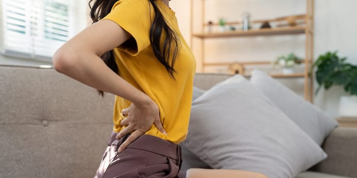 Young woman in a yellow shirt holding her lower back in pain while sitting on a couch, indicating physical discomfort