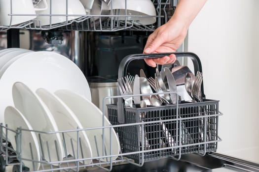 Female hand unloading dishwasher with tableware. Kitchen appliances, lifestyle view. Woman takes a cutlery basket from the dishwasher. Concept of modern technologies in the kitchen