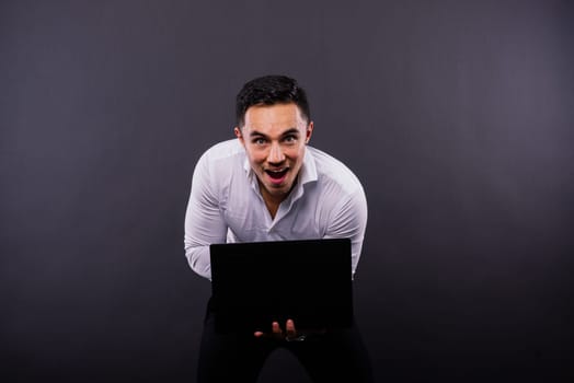 Image of a young cheerful businessman holding and using laptop isolated over dark background