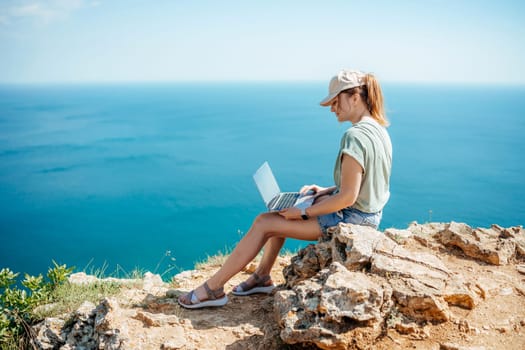 A woman is sitting on a rock by the ocean, using her laptop. Concept of relaxation and tranquility, as the woman enjoys the beautiful view of the ocean while working on her laptop