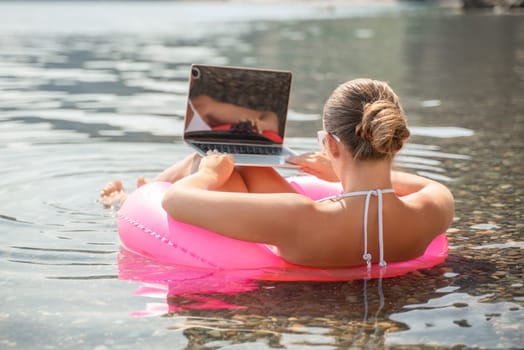 A woman is sitting on a pink inflatable raft in the water, using a laptop. Concept of relaxation and leisure, as the woman is enjoying her time in the water while working on her laptop