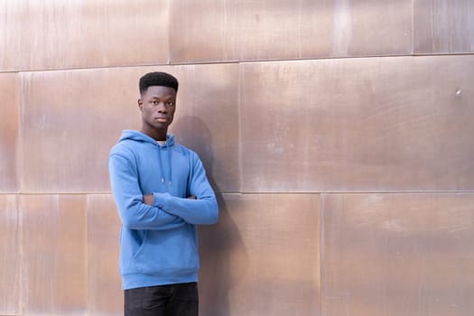 A young man in a blue hoodie stands in front of a wall. He is looking at the camera with his arms crossed. Scene is serious and contemplative