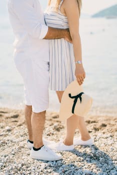 Man hugs woman with a straw hat in her hand while standing on the seashore. Cropped. Faceless. High quality photo