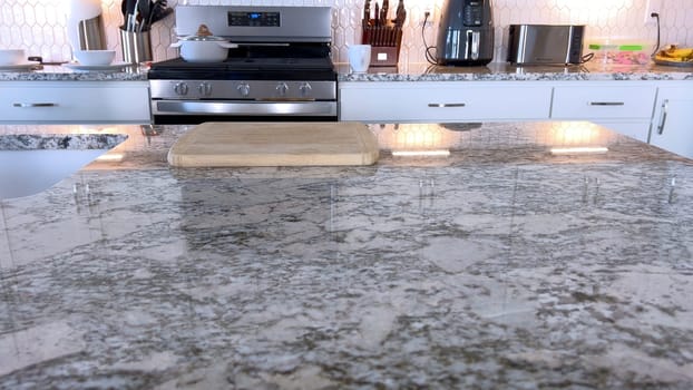 A beautifully designed modern kitchen featuring polished granite countertops and stainless steel appliances. The countertop is clean and organized, with a wooden cutting board placed in the center, ready for meal preparation. The backsplash has a white hexagonal tile pattern, adding a touch of elegance to the space.