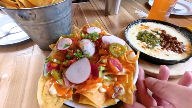 A mouthwatering plate of loaded nachos topped with cheese, radishes, jalapenos, and fresh vegetables. Accompanied by a bucket of tortilla chips and a skillet of creamy dip with peppers, perfect for a tasty dining experience.