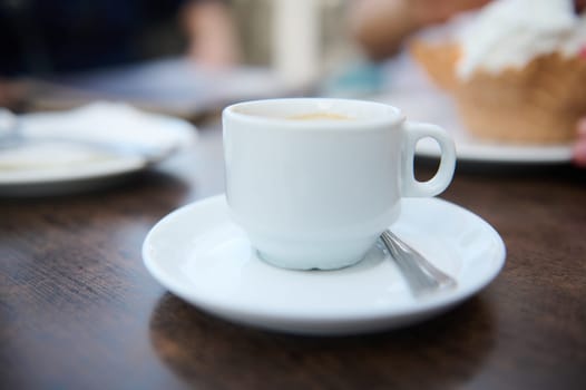 Close-up image of a white coffee cup on a saucer with a spoon, positioned on a wooden table in a cafe setting.