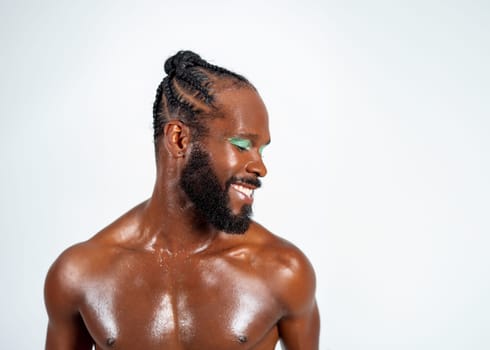 Portrait of shirtless muscular African American gay man with green eyeshadow. Confident bearded man smiling. Happy gay with bright makeup on white background.