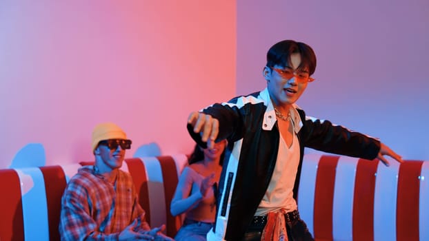 Asian man standing and dancing with pop music while multicultural friend sitting behind at home with led light. Attractive street dancer moving to hip hop music while looking at camera. Regalement.