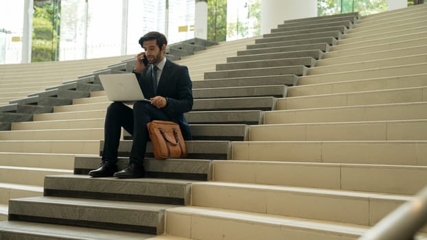 Skilled investor working or planing strategy by using laptop at stair. Professional business man wearing suit while working and typing data analysis by using laptop and talking to phone. Exultant.