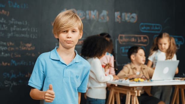 Smart boy showing thumb while diverse friend working or learning engineering code or prompt in STEM technology classroom. Caucasian student standing at camera while children using laptop. Erudition.