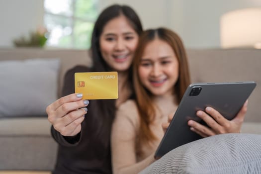 A happy lesbian couple holding credit cards while shopping online using a tablet, showcasing modern e-commerce and technology in a comfortable home setting.