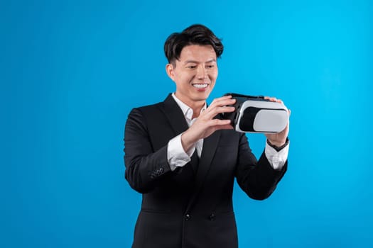 Smiling Asian businessman looking through VR connecting financial business report futuristic metaverse data analytics power bi technology virtual reality meta isolated blue copyspace. Contrivance.