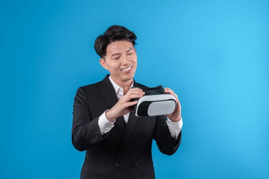 Smiling Asian businessman looking through VR connecting financial business report futuristic metaverse data analytics power bi technology virtual reality meta isolated blue copyspace. Contrivance.