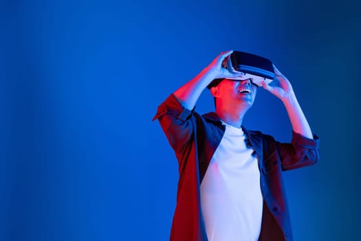 Asian man looking through VR enjoy watching fantastic meta on interesting object technology blue neon lighting background on virtual reality metaverse new world in 3D hologram innovation. Contrivance.