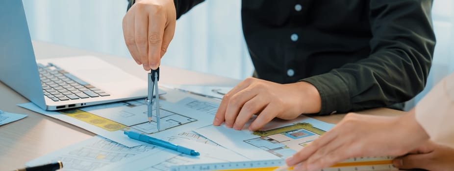 A cropped image of professional engineer using divider to measure blueprint at meeting table with blueprint, laptop and architectural equipment scatter around. Closeup. Delineation.